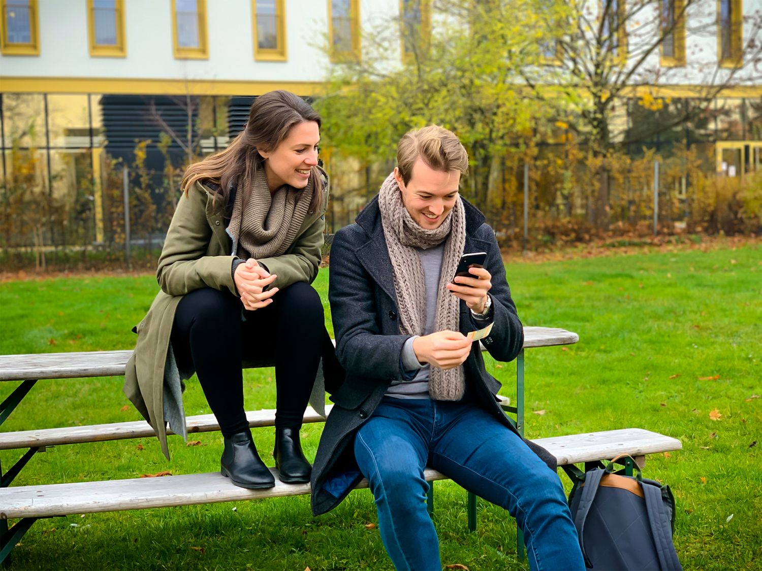 Zwei junge Menschen sitzen auf einer Bank im Grünen und schauen auf ein Smartphone.